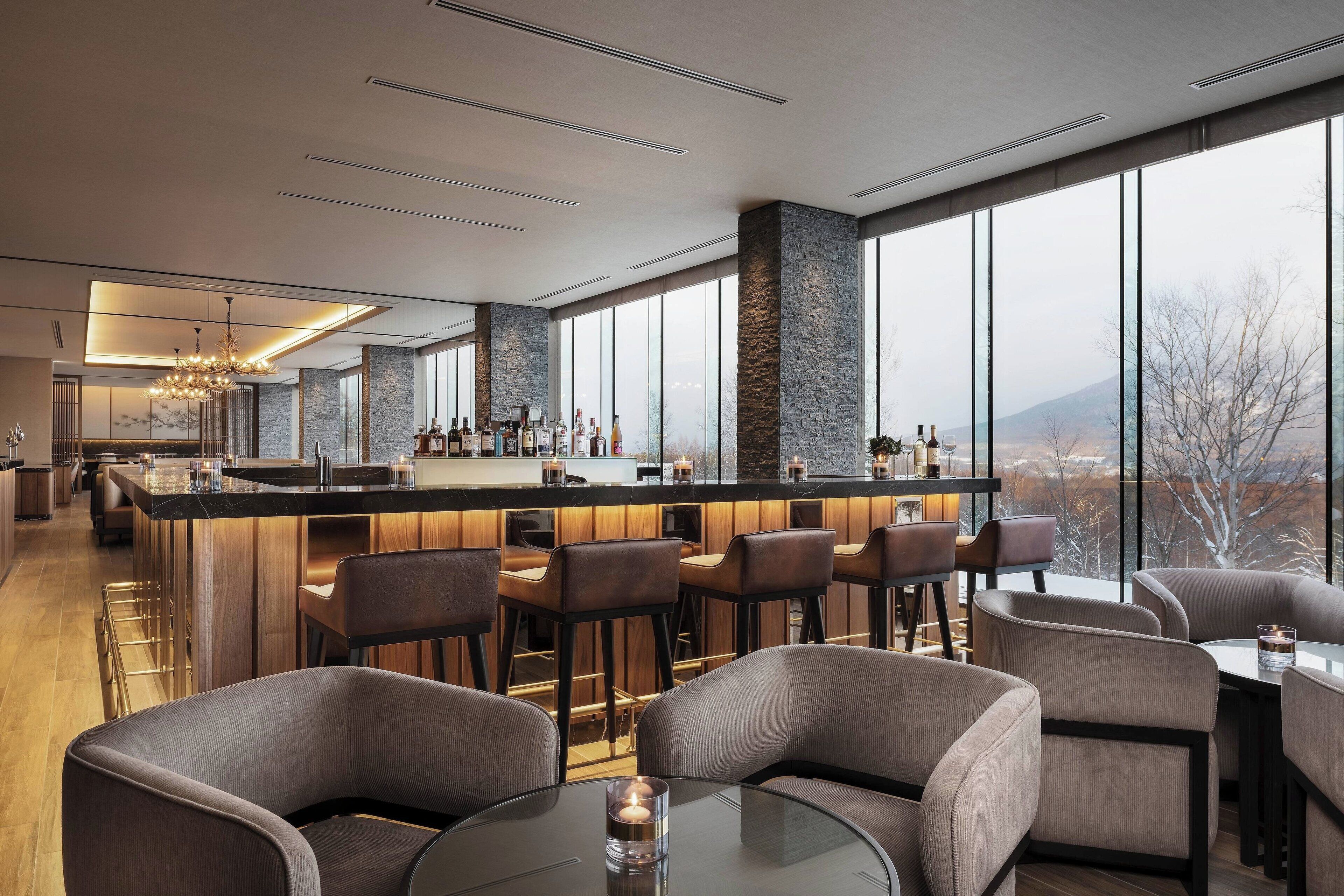 A modern bar area with a high bar counter, seating with bar stools, and large windows offering a view of the outdoors.