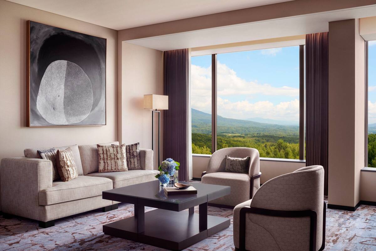 A modern hotel room with a sofa, armchair, and a large window offering a mountain view.