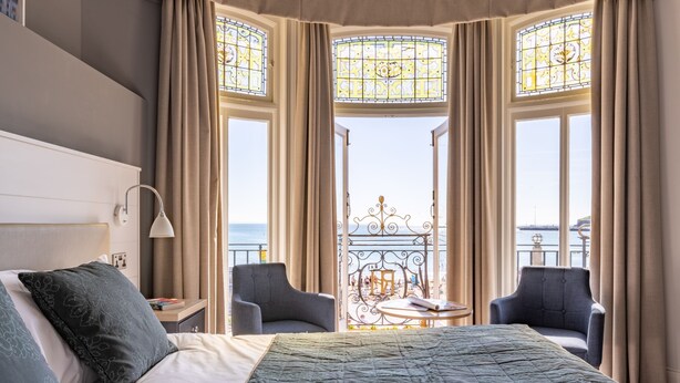 A hotel room with a large bed, two armchairs, a small table, and a view of the sea through stained glass windows. - The Royal Hotel (Weymouth)