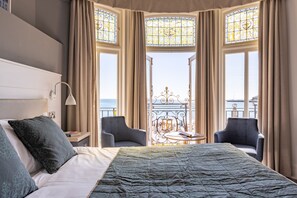 A hotel room with a large bed, two armchairs, a small table, and a view of the sea through stained glass windows.