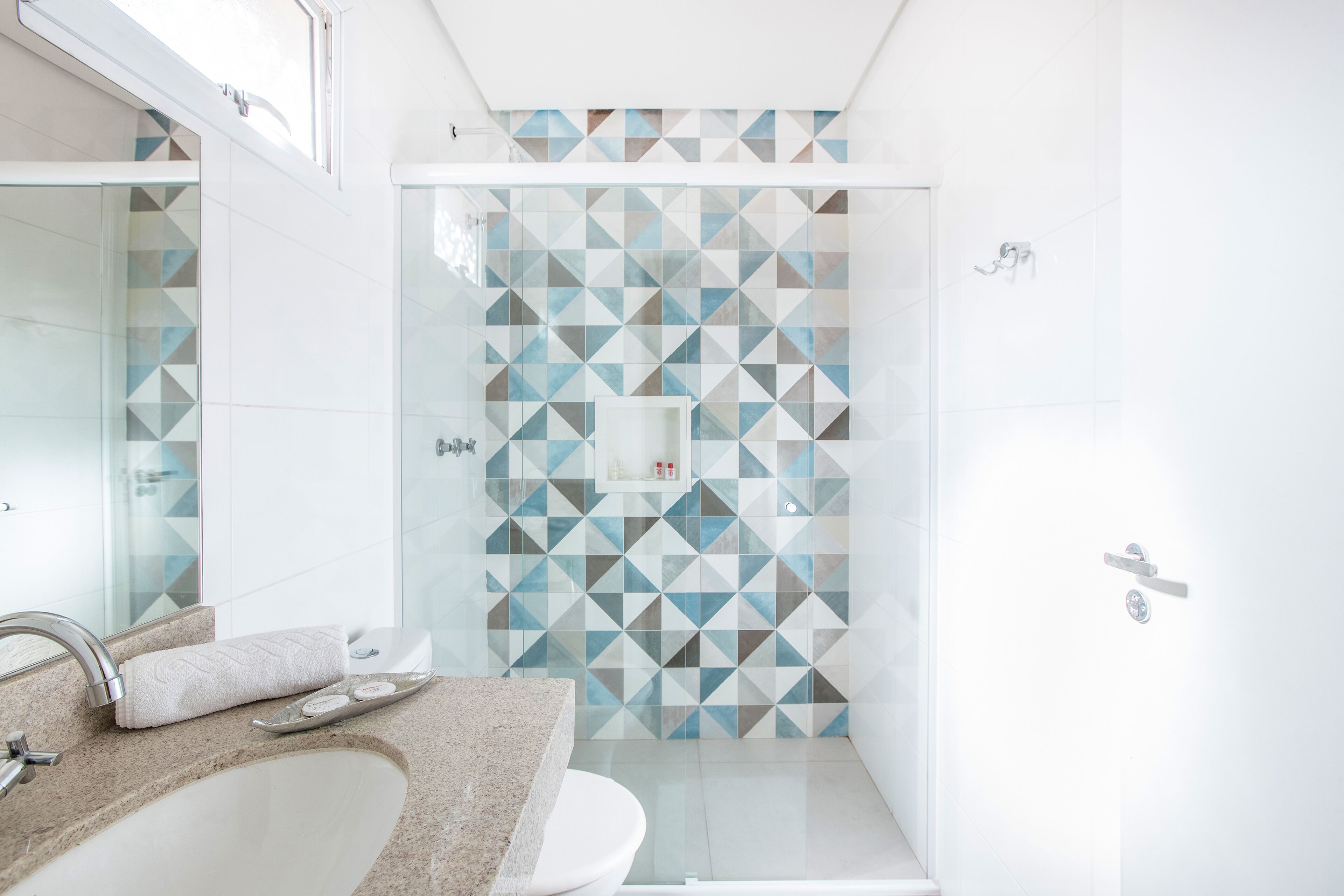 Et moderne badeværelse med et geometrisk flisemønster bag toilettet, en brusekabine i glas og et spejl med vask.