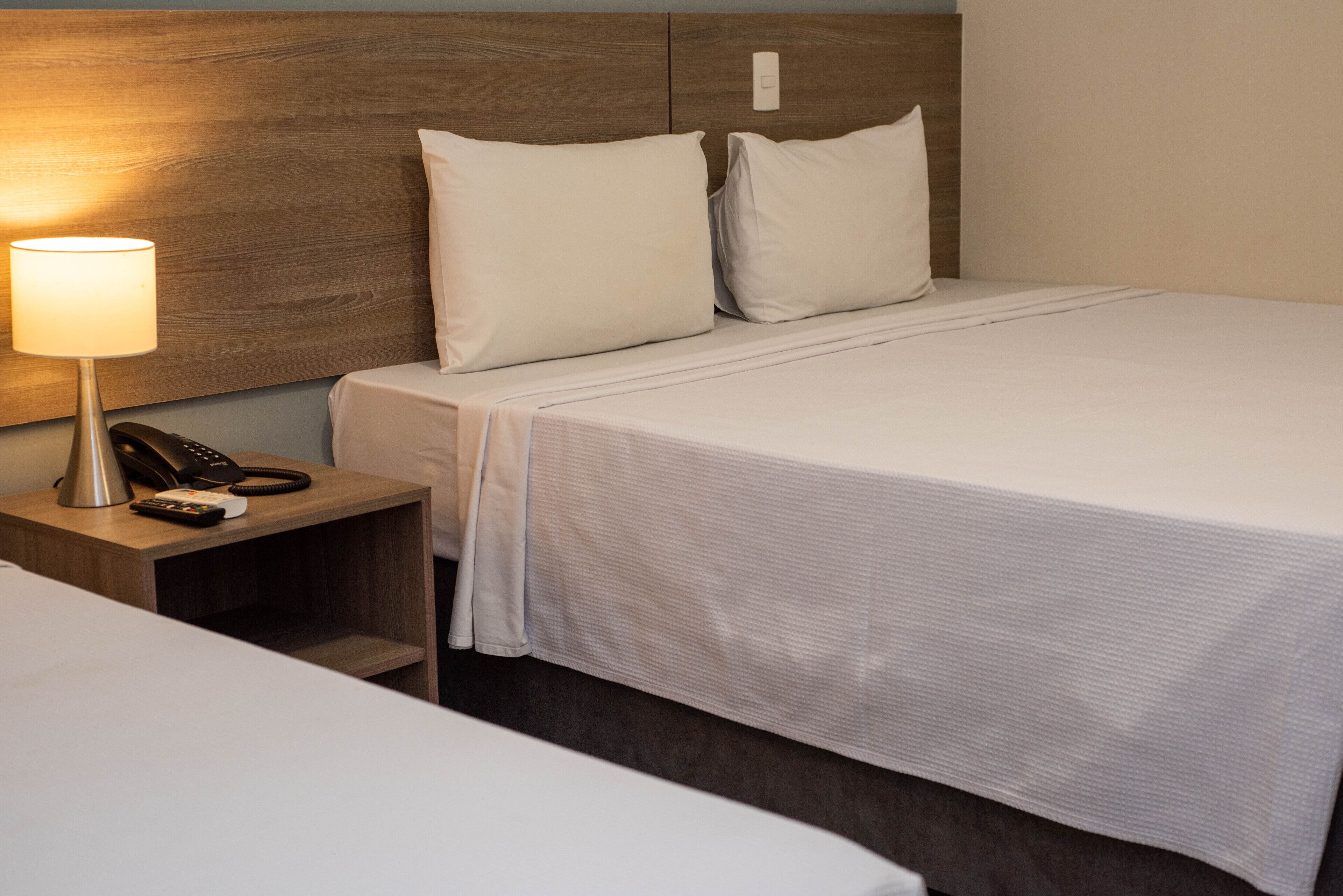 Ein Hotelzimmer mit einem ordentlich bezogenen Bett, einer Nachttischlampe und einem Telefon auf einem Holznachttisch.