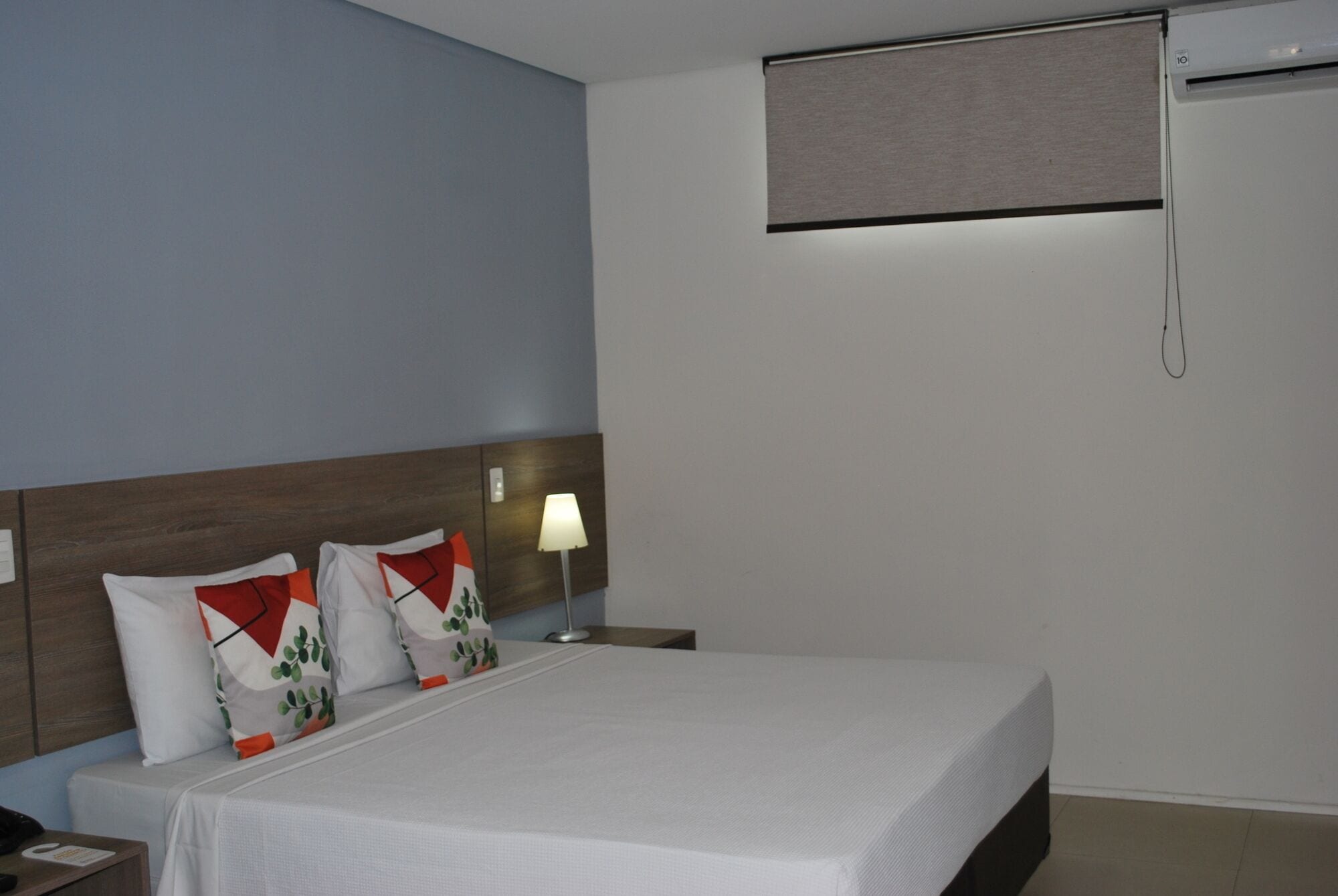 A hotel room with a bed, a nightstand, a lamp, and a window with a blind.