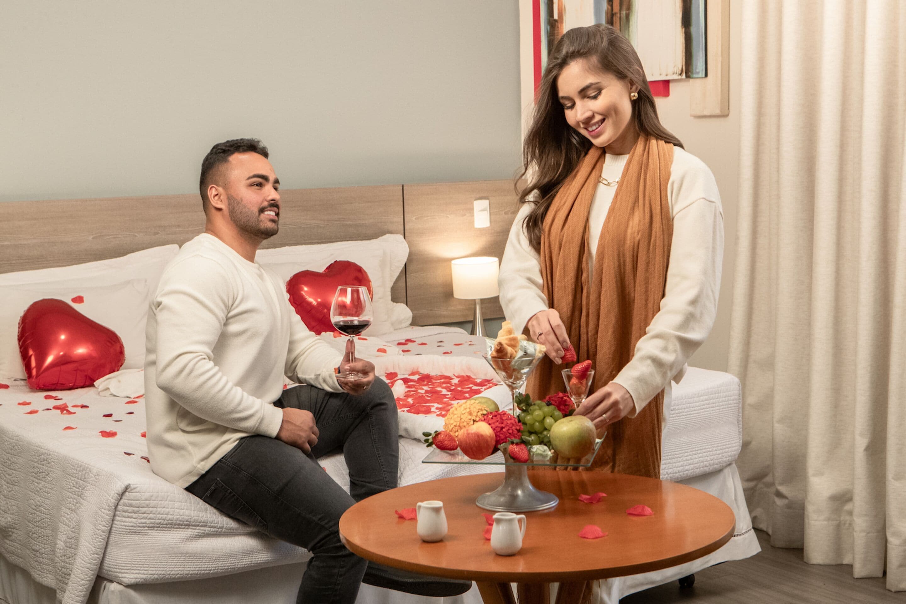 A couple sitting on a bed with heart-shaped pillows, a table with fruit and a wine glass, and a bedside lamp.