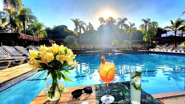 Un cadre agréable au bord de la piscine, avec des chaises longues, une table où sont disposées des boissons et des fleurs, et des palmiers en arrière-plan.