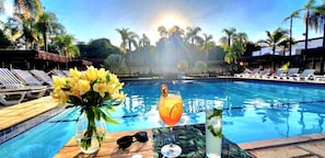 Un ambiente junto a la piscina con sillones de descanso, una mesa con bebidas y flores, y palmeras al fondo.