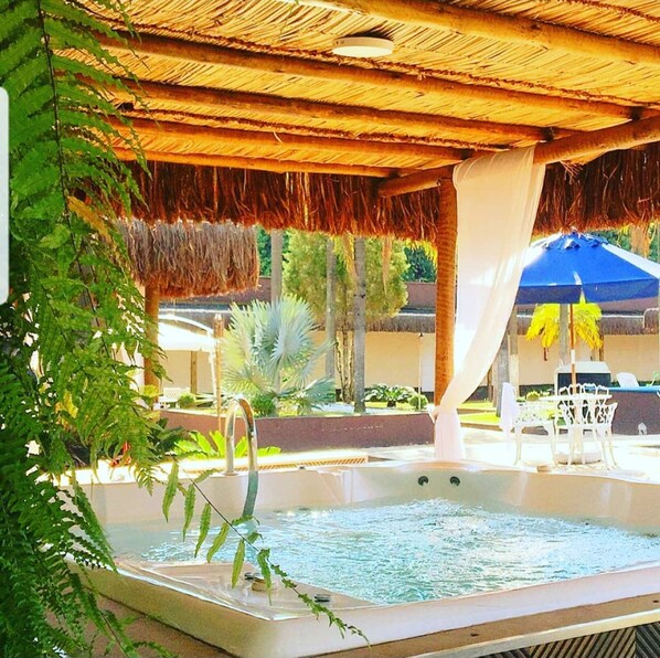 Zona de hidromasaje con techo de paja, rodeada de follaje y un ambiente junto a la piscina.