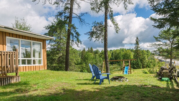 A wooden cabin with a large window, surrounded by a grassy yard and tall trees. - C1 Spacious Cottage - Nature Getaway in Peaceful Northern Ontario (Cobalt)