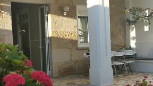 Ein Steinhaus mit einer Terrassenfläche, die einen Tisch und Stühle, eine Tür sowie ein Fenster mit Vorhang zeigt.