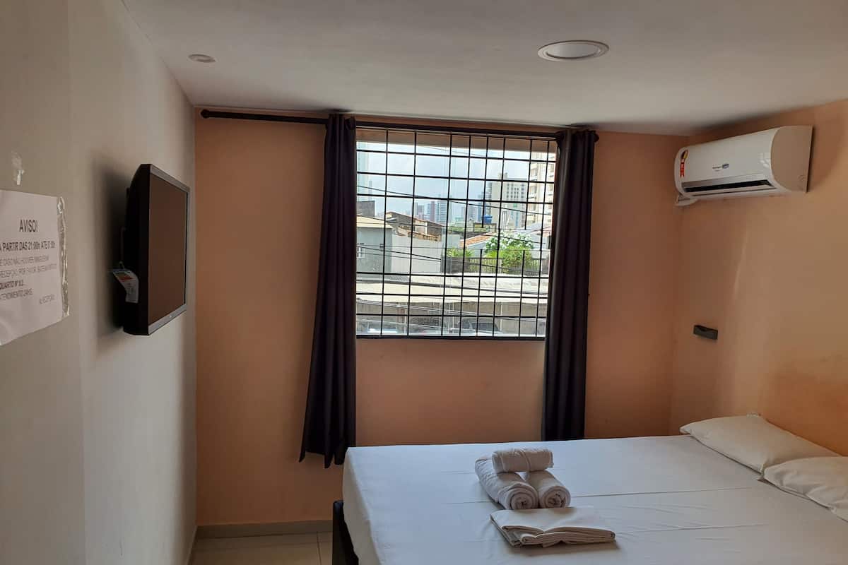 A hotel room with a bed, a window with curtains, an air conditioner, a TV mounted on the wall, and a notice on the wall.