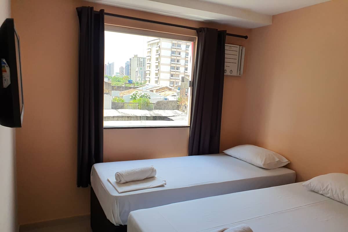 A hotel room with two beds, a window with curtains, a wall-mounted air conditioner, and a view of buildings outside.
