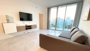A modern living room with a large flat-screen TV mounted on the wall, a built-in entertainment unit, a large window with a city view, and a comfortable sofa. - Sunny Isles Penthouse Right Near the Beach (Sunny Isles)