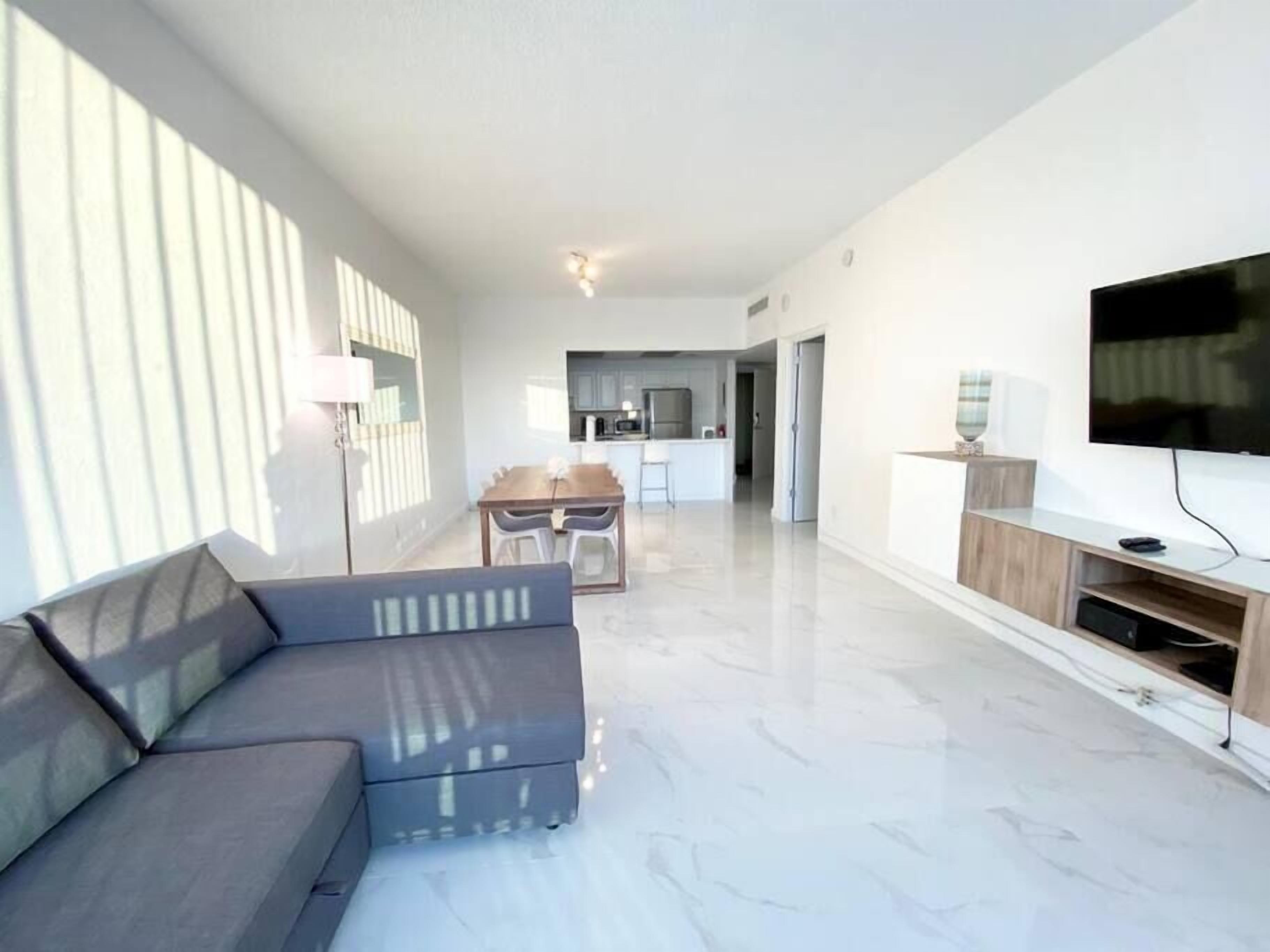 A modern living room with a grey sofa, a flat-screen TV on a wooden stand, and a kitchen in the background.