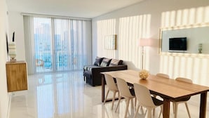 A modern living room with a dining area, large windows with sheer curtains, a flat-screen TV, and a city view. - Sunny Isles Penthouse Right Near the Beach (Sunny Isles)