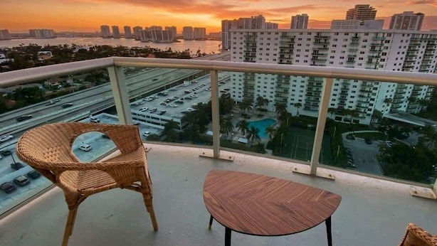 A balcony with a wicker chair and a small table, overlooking a cityscape with high-rise buildings and a body of water during sunset. - Sunny Isles Penthouse Right Near the Beach (Sunny Isles)