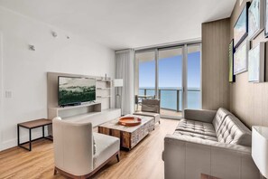 Una sala de estar moderna con un televisor de pantalla plana grande, una mesa de centro de madera, un sofá color beige y vistas al océano a través de ventanas amplias.