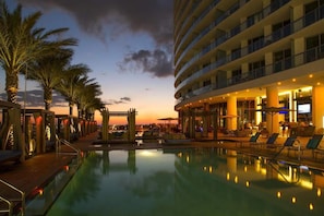 A hotel pool area with a sunset view, palm trees, and a multi-story building with balconies. - Luxurious Ocean View 2 Bedroom Escape (Hollywood)