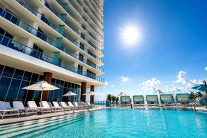 A hotel pool area with sun loungers and umbrellas, a clear blue sky, and a modern building with balconies. - Luxurious Ocean View 2 Bedroom Escape (Hollywood)