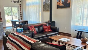 Un salon avec un canapé en cuir noir, une table basse en bois, un plaid à rayures, une cheminée et un coin repas comprenant une table et des chaises.