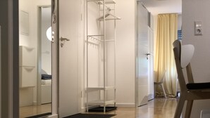 Moderni hotellihuone, jossa on puulattia, valkoinen vaatekaappi, lasiovi, joka johtaa kylpyhuoneeseen, sekä pieni oleskelualue, jossa on tuoli ja pöytä.