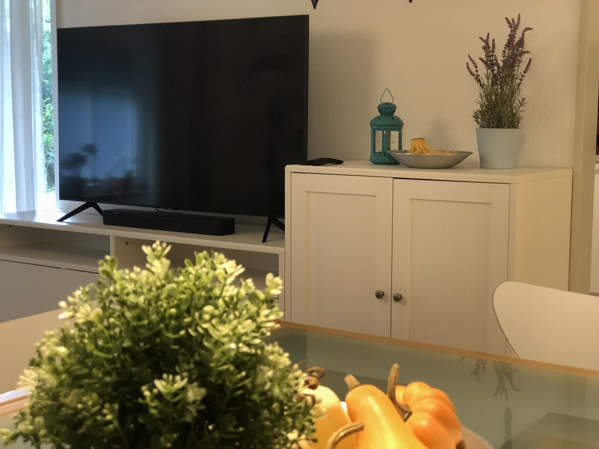 Una sala de estar moderna con un televisor de pantalla plana montado en la pared, un mueble blanco con puertas, un farolillo azul turquesa y una planta en maceta.