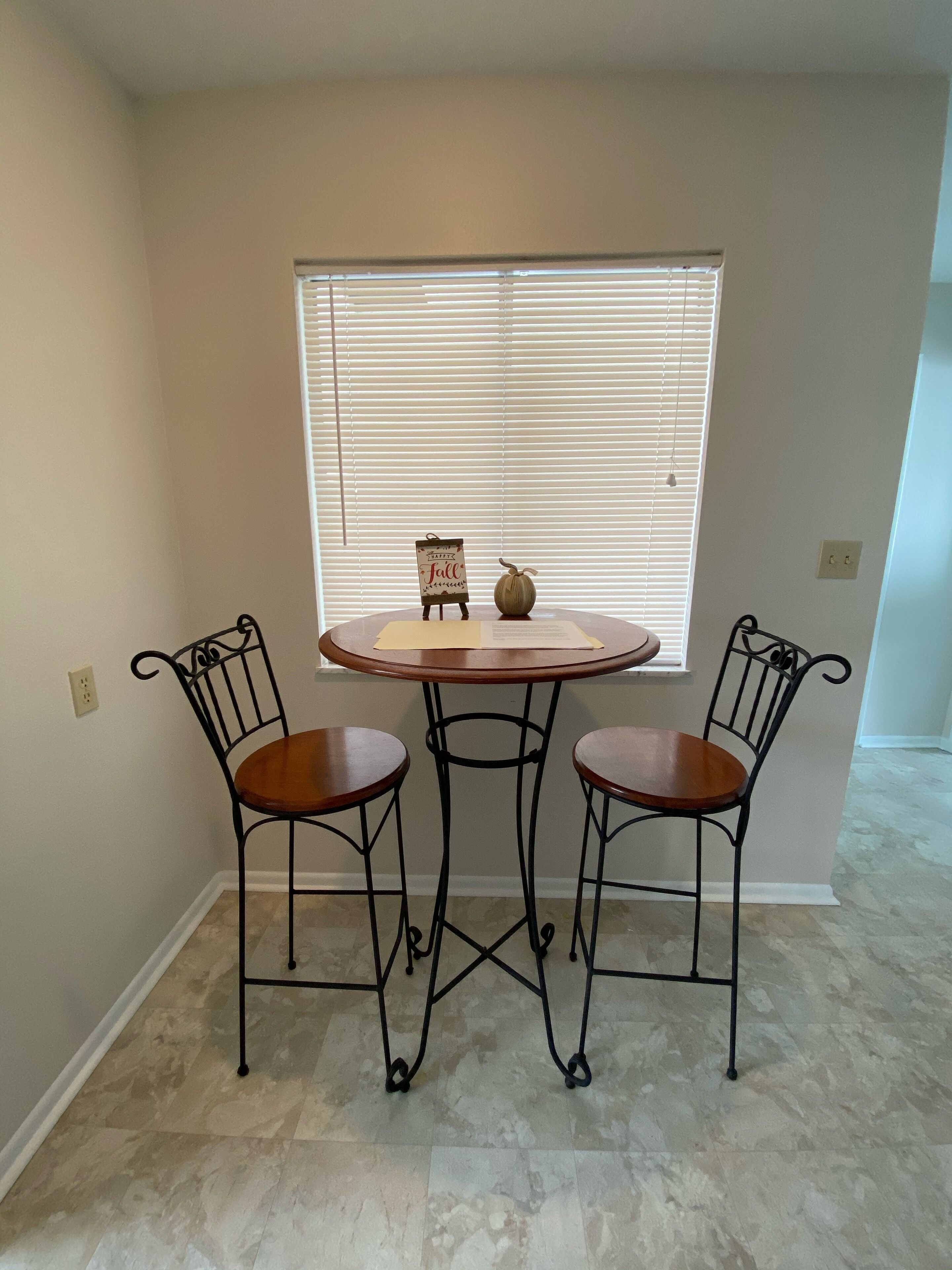A dining area with a round table and two chairs, a small decorative item on the table, and a window with blinds in the background.