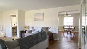 A spacious living room with a grey sofa, wooden flooring, a dining area with a table and chairs, and a ceiling fan.