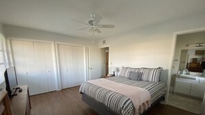 A bedroom with a bed, a ceiling fan, a desk with a computer, and a bathroom with a sink and mirror.