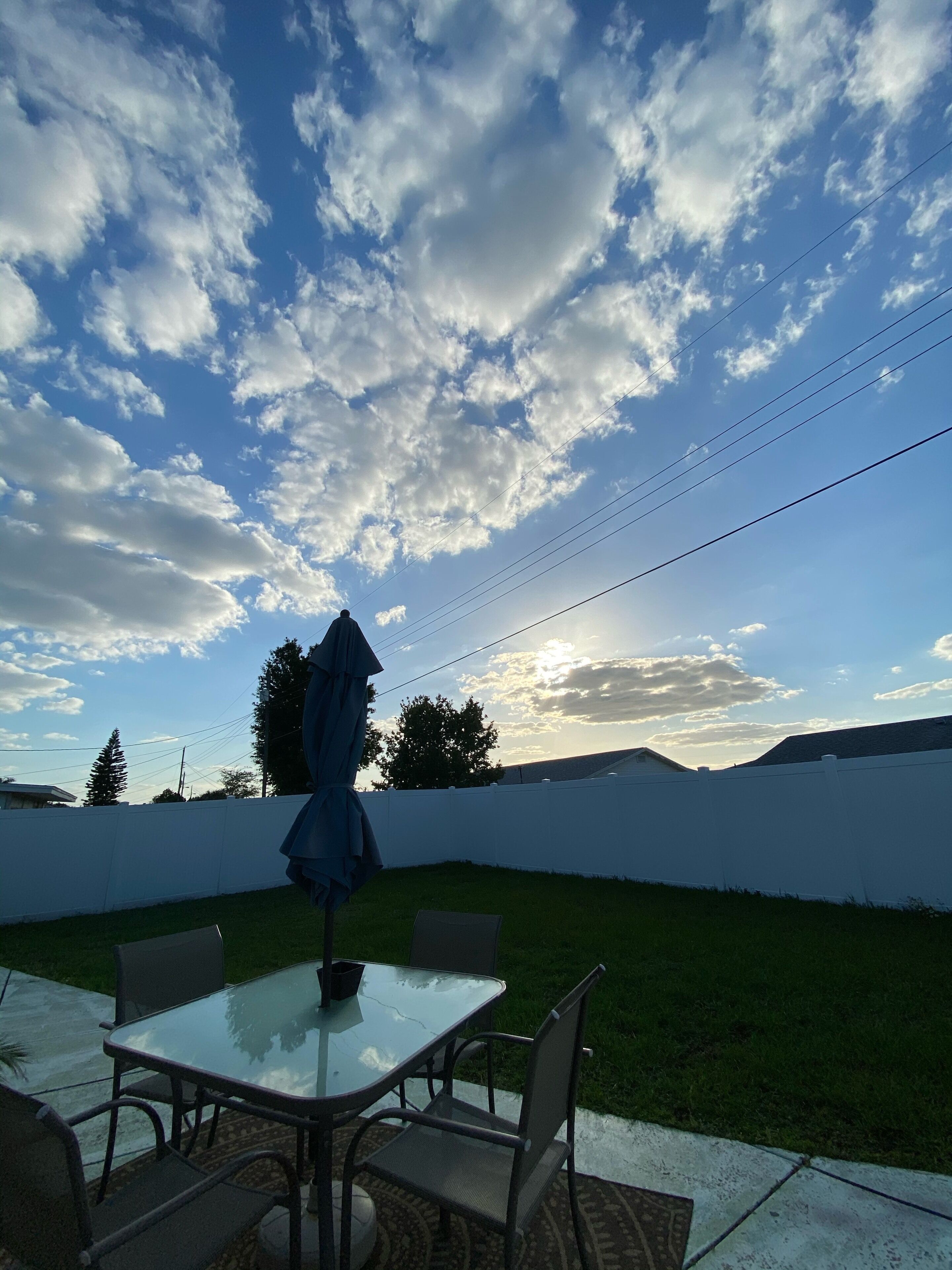 A glass table with chairs and an umbrella in a backyard with a white fence and a grassy lawn.