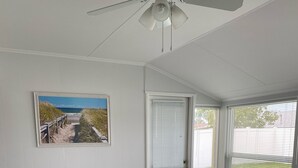 A room with a beige sofa, a ceiling fan, a door with blinds, a framed picture of a beach, and a potted plant.