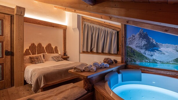 A wooden room with a bed, a window with curtains, a painting of mountains, and a bathtub. - Ciandolada 2 Wellness (Vodo Cadore)