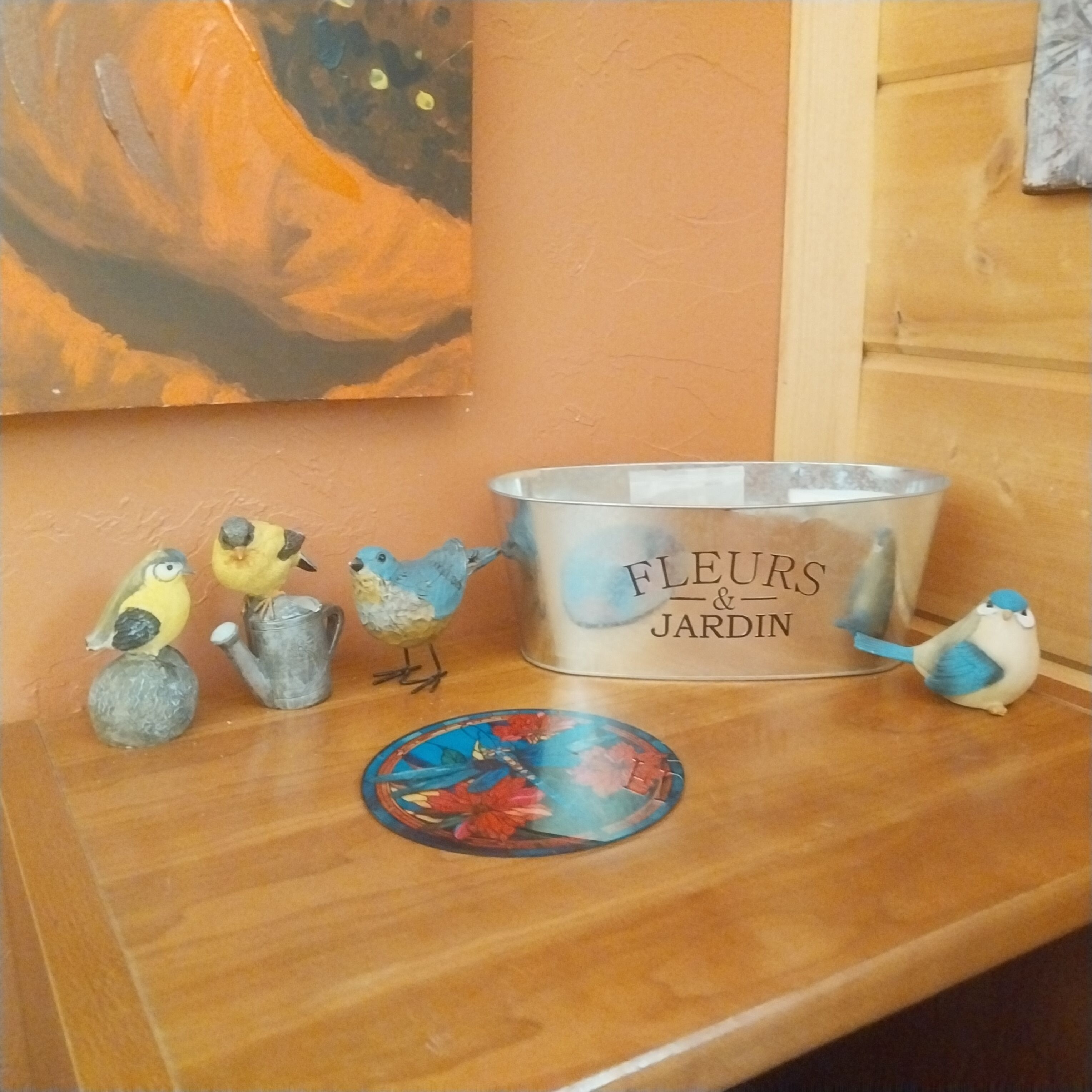 Ein Holztisch mit einem Metallbehälter mit der Aufschrift "FLEURS & JARDIN“, dekorativen Vogelbildern und einem floralen Untersetzer.