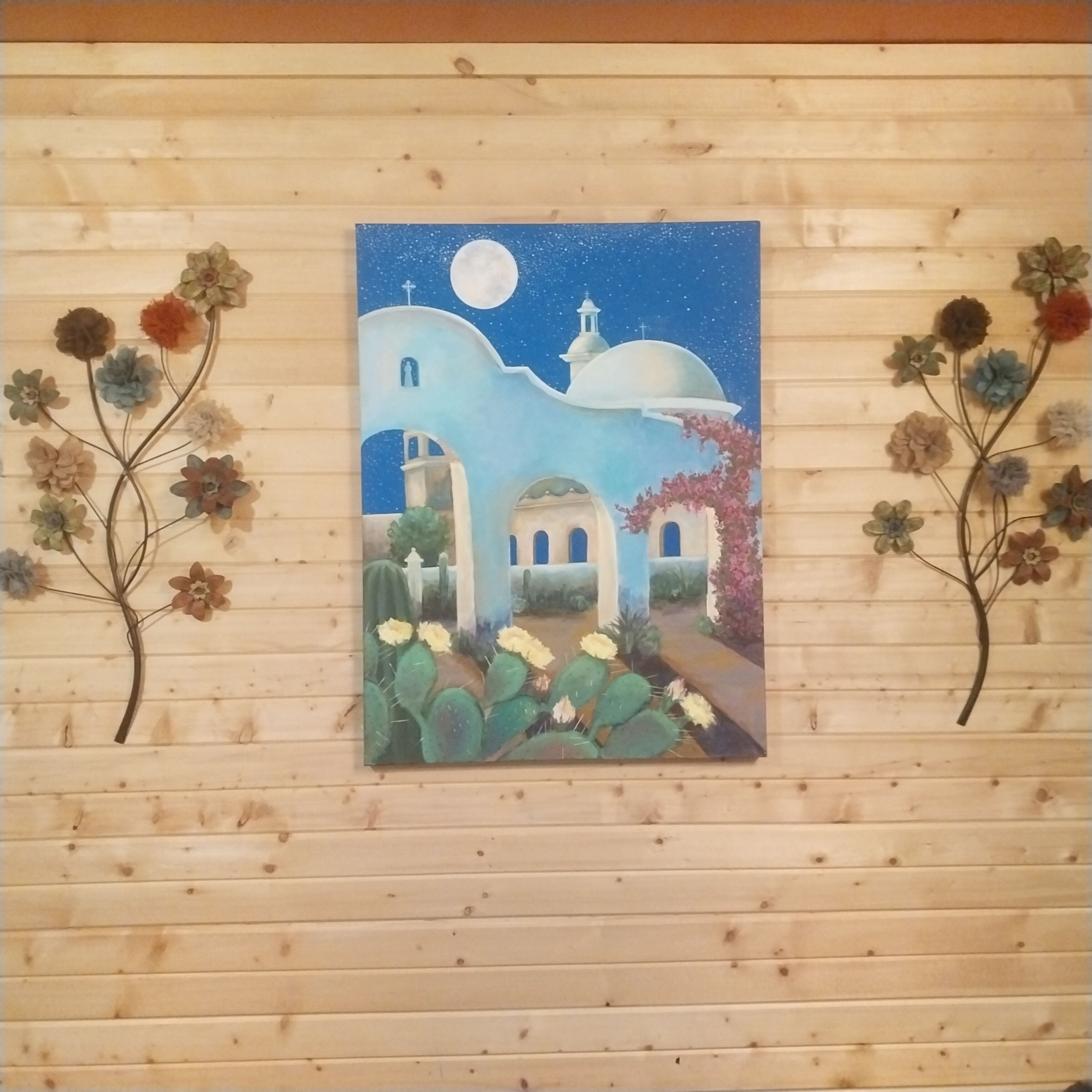 Una pintura de una escena nocturna con una luna, una iglesia y cactus en una pared de madera.