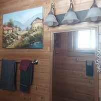 A wooden cabin interior with a painting of a rural scene, a door with a towel rack, and a window.