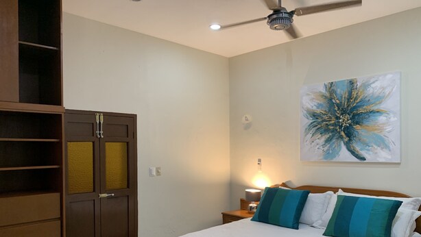 Un dormitorio con cama, mesita de noche, lámpara, ventilador de techo y un cuadro abstracto de gran tamaño en la pared.