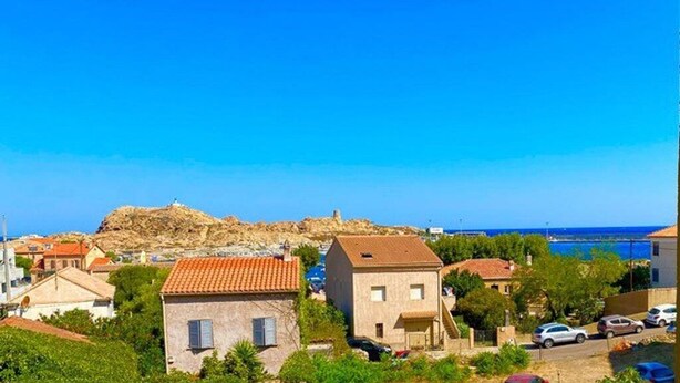 Uma vila costeira com edifícios de pedra, uma colina rochosa e um céu azul límpido.