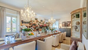 A spacious dining area with a long wooden table, white upholstered chairs, and a chandelier.