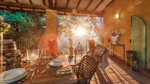 An outdoor dining area with a wooden table set for two, surrounded by wicker chairs, and a view of the sunset through trees.