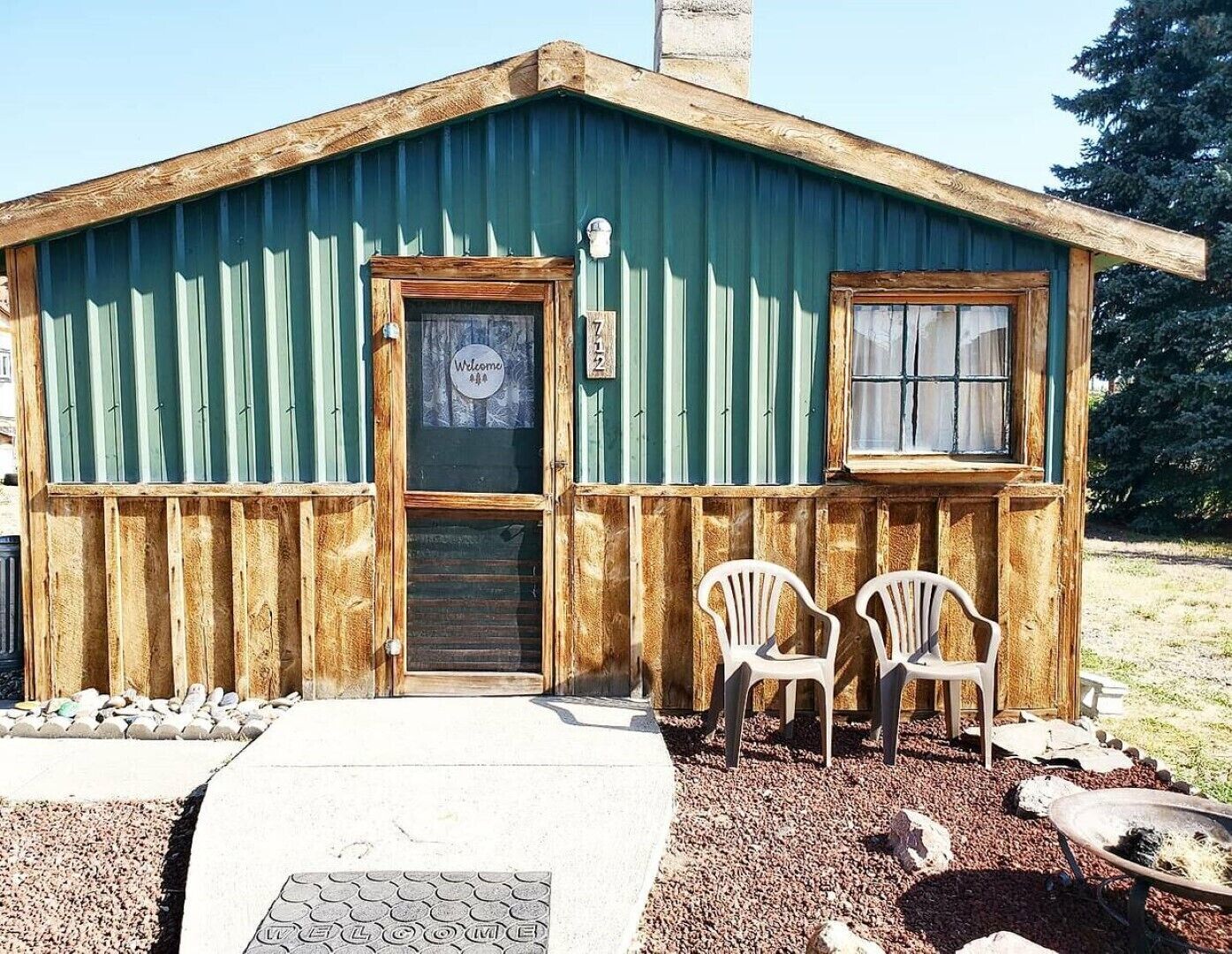 A rustic cabin with a wooden exterior, a welcome sign, and two plastic chairs outside.