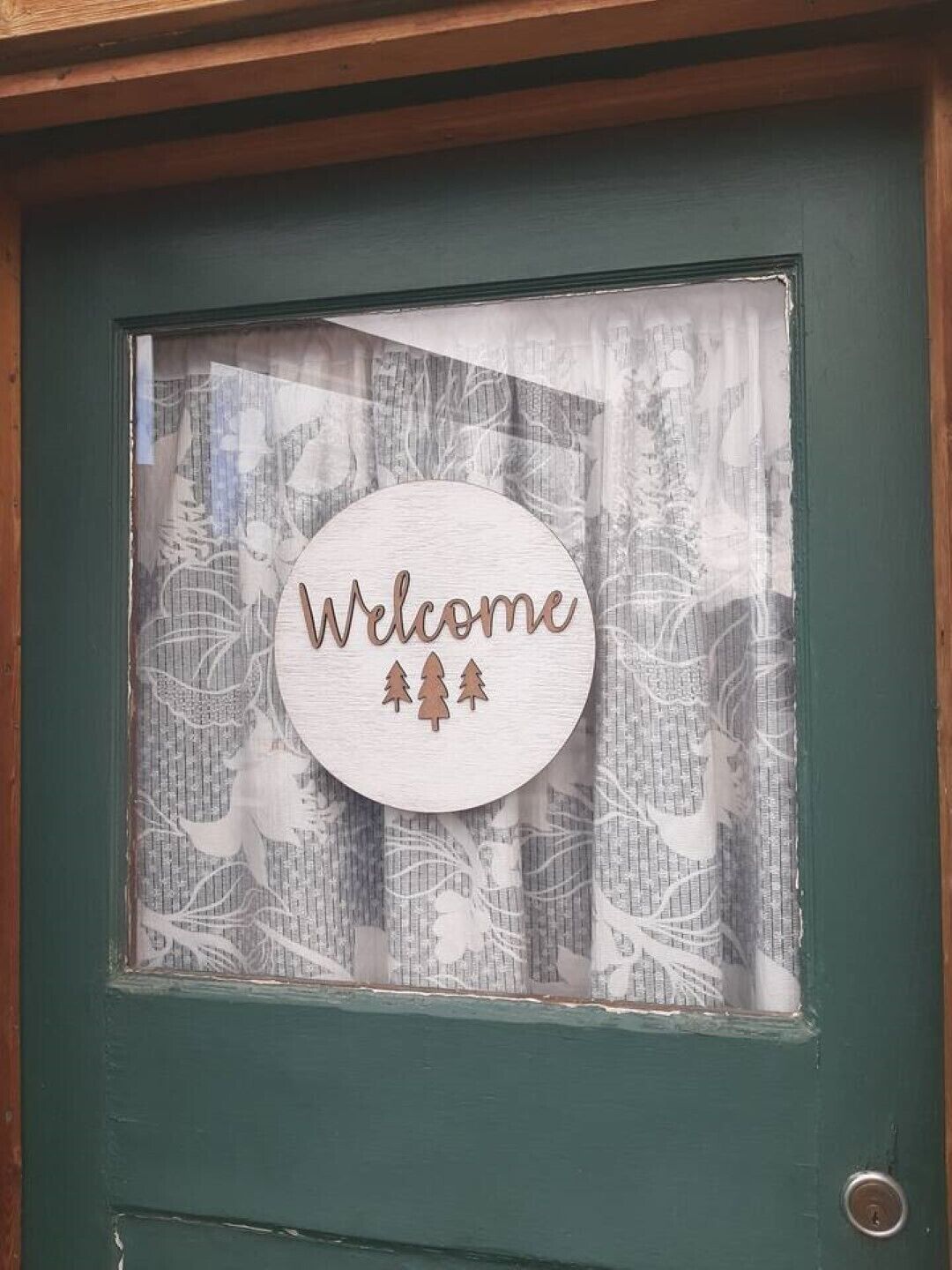 A green door with a window featuring a 'Welcome' sign and three pine tree silhouettes.