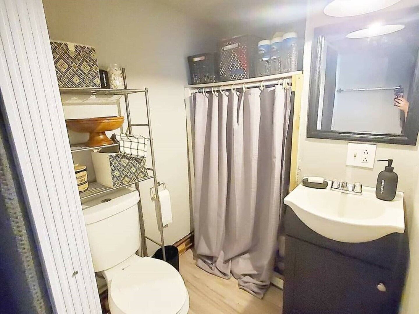 A compact bathroom with a toilet, a sink, a shelf with toiletries, and a shower curtain.