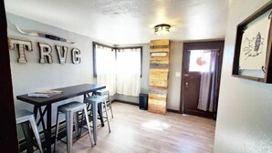 A dining area with a wooden table and bar stools, a wall-mounted sign reading "TRVC", and a wooden accent wall. - The Rustic Valley Cabin (Challis)
