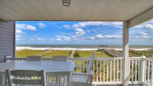 A covered outdoor area with a table and chairs, overlooking a beach with a boardwalk and ocean view.