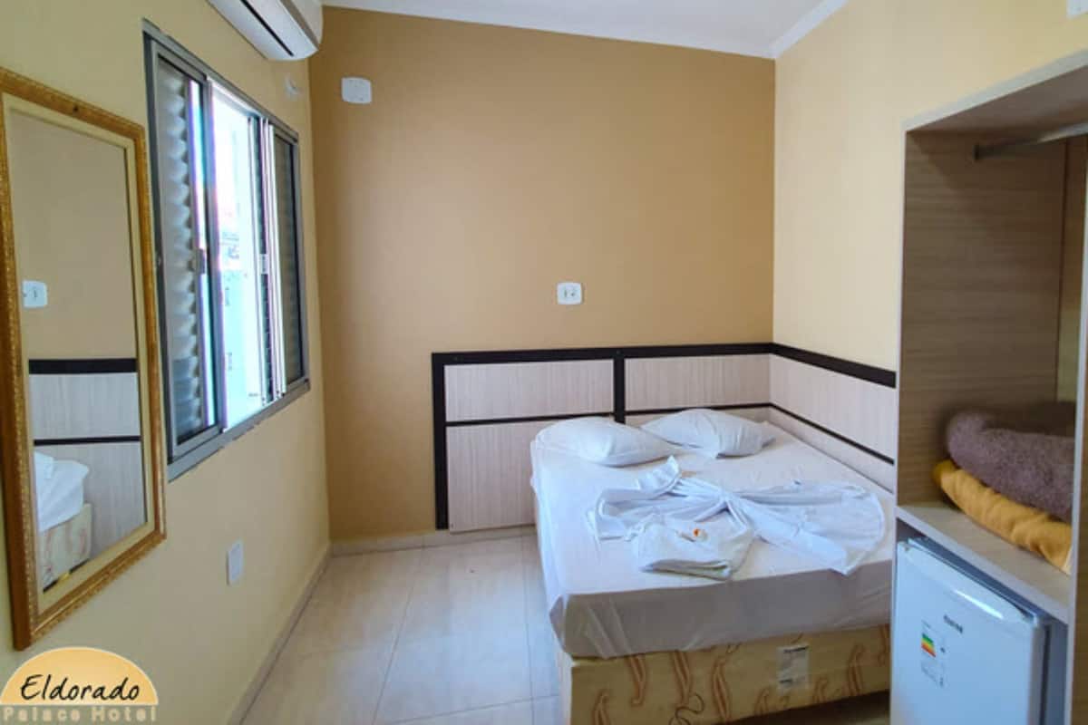 A hotel room with a bed, a mirror, an air conditioner, and a small refrigerator.