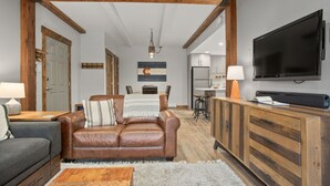 Een woonkamer met een bruine leren bank, een grijze zithoek, houten balken, een televisie, en een keuken met kasten en een koelkast.