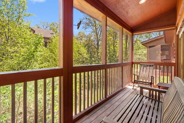 Eine Holzterrasse mit Bank und Tisch, umgeben von einem Geländer, mit Blick auf einen bewaldeten Bereich.