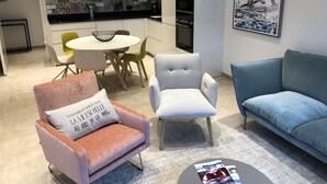Un salon moderne avec un fauteuil rose, un fauteuil gris et un canapé bleu. On y trouve également un coin repas avec une table et des chaises blanches, et une cuisine à l’arrière-plan.