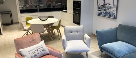 A modern living room with a pink armchair, a grey armchair, and a blue sofa. There is a dining area with a white table and chairs, and a kitchen in the background.