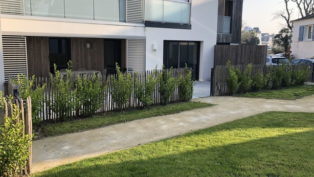 Un immeuble résidentiel moderne à deux étages, doté d’un espace extérieur couvert avec des sièges, d’une clôture en bois et d’une pelouse bien entretenue.