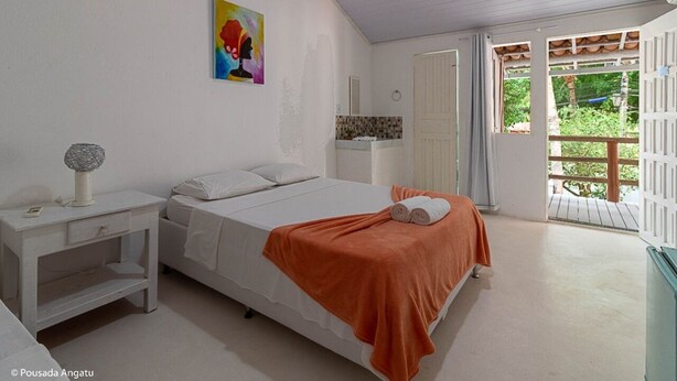 A bedroom with a bed, a nightstand, a painting, a bathroom with a mirror, and a balcony with a view of greenery.
